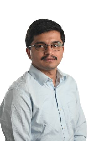 serious person with dark hair and moustache wearing spectacles and light grey shirt