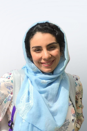 smiling person with dark hair and light blue head scarf wearing a floral print on cream top